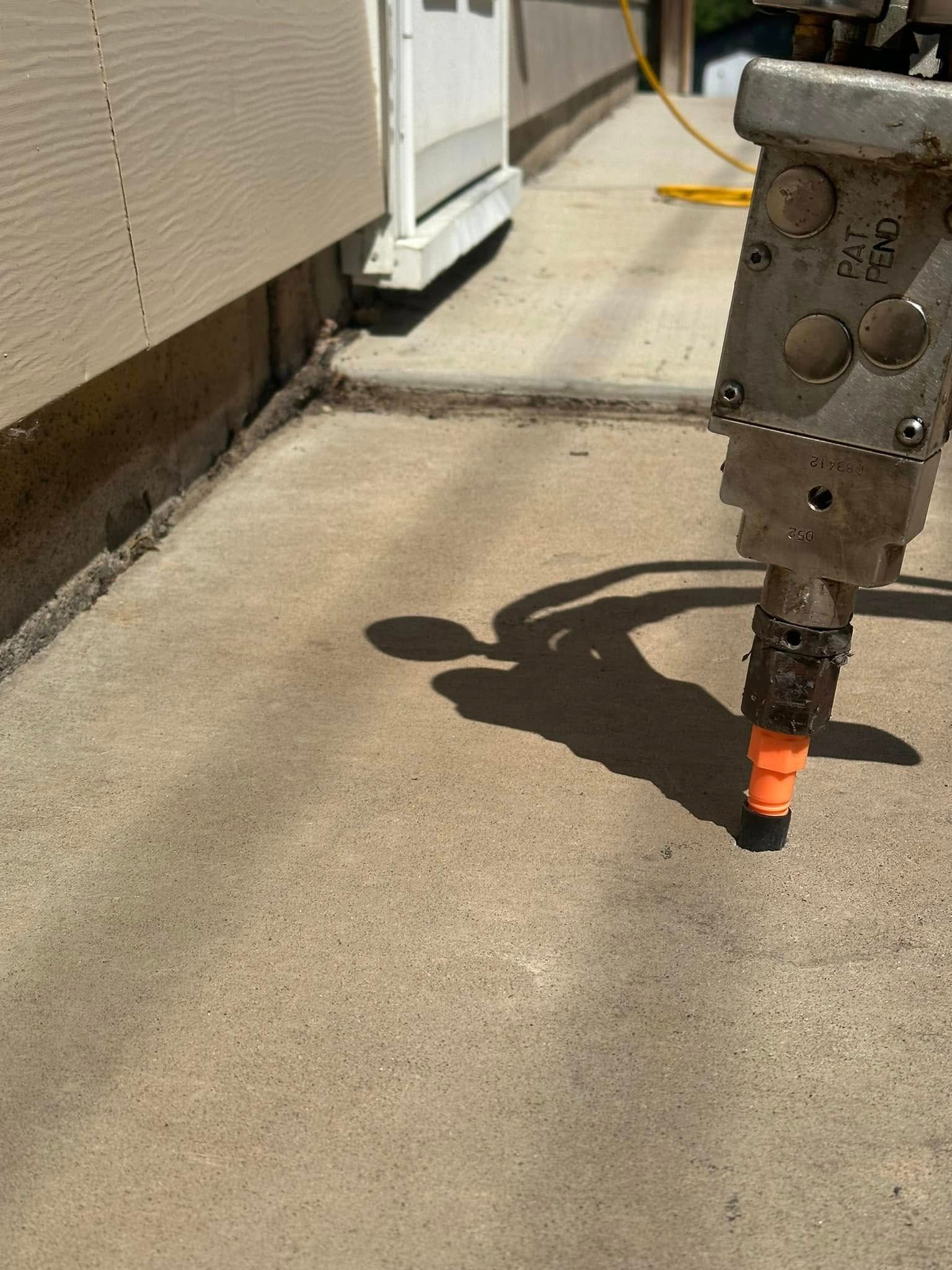 A tool working on a concrete surface next to a building. The tool has a visible orange nozzle.