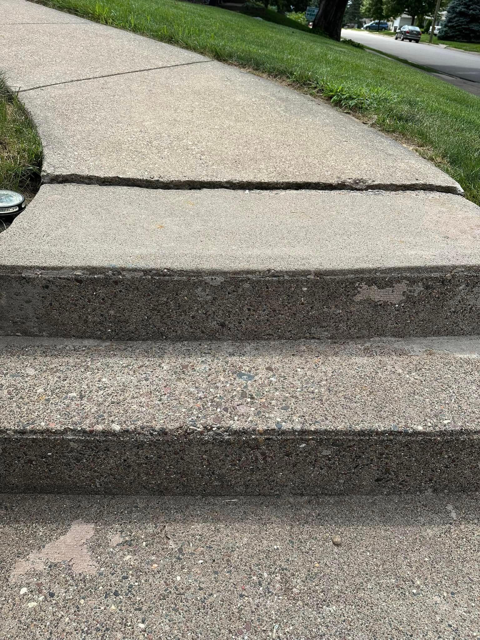 Concrete steps leading up to a sidewalk, with cracks and wear. Green grass borders the path.