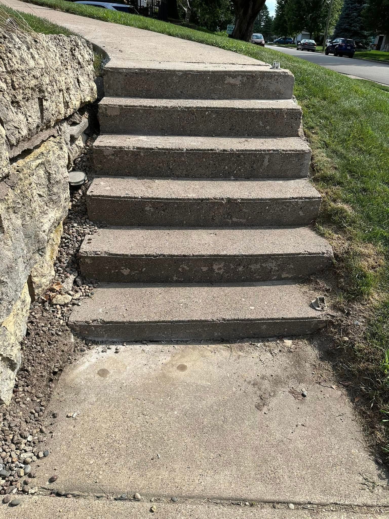 Concrete stairs leading up from a sidewalk, next to a stone retaining wall and grass.