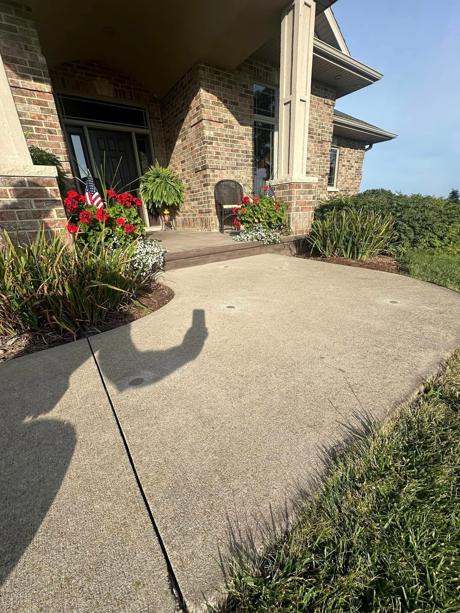 Concrete walkway leads to a brick house entrance with landscaping. Sunny day with shadows.
