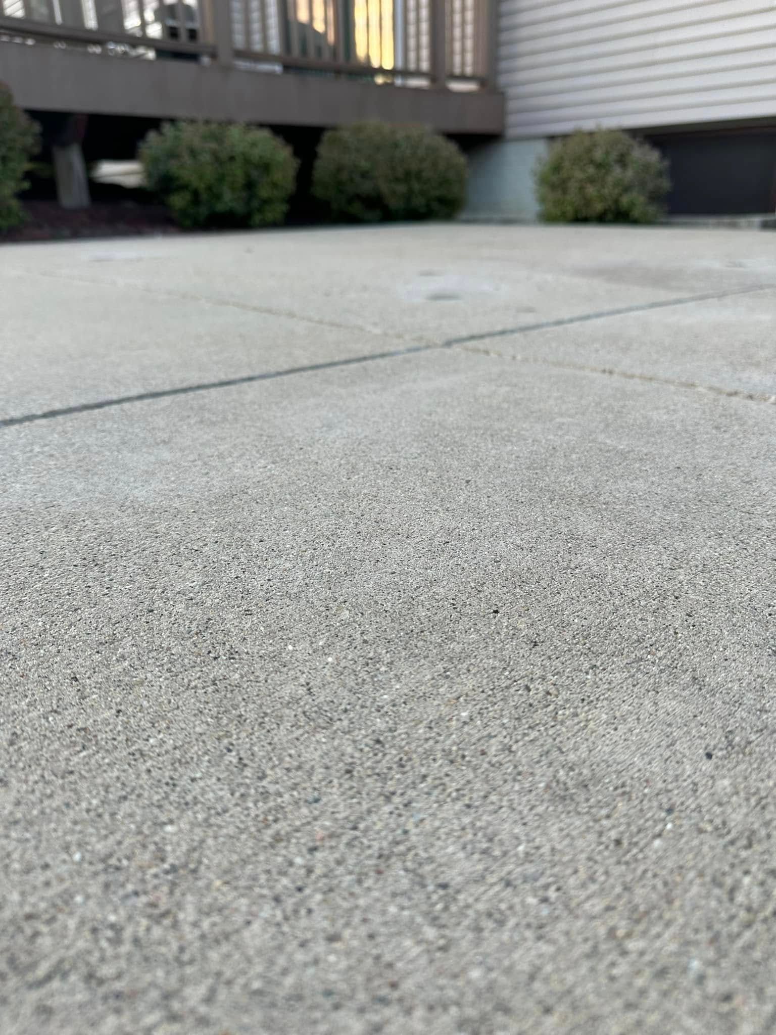 Close-up view of a textured concrete patio with a deck and shrubbery in the background.