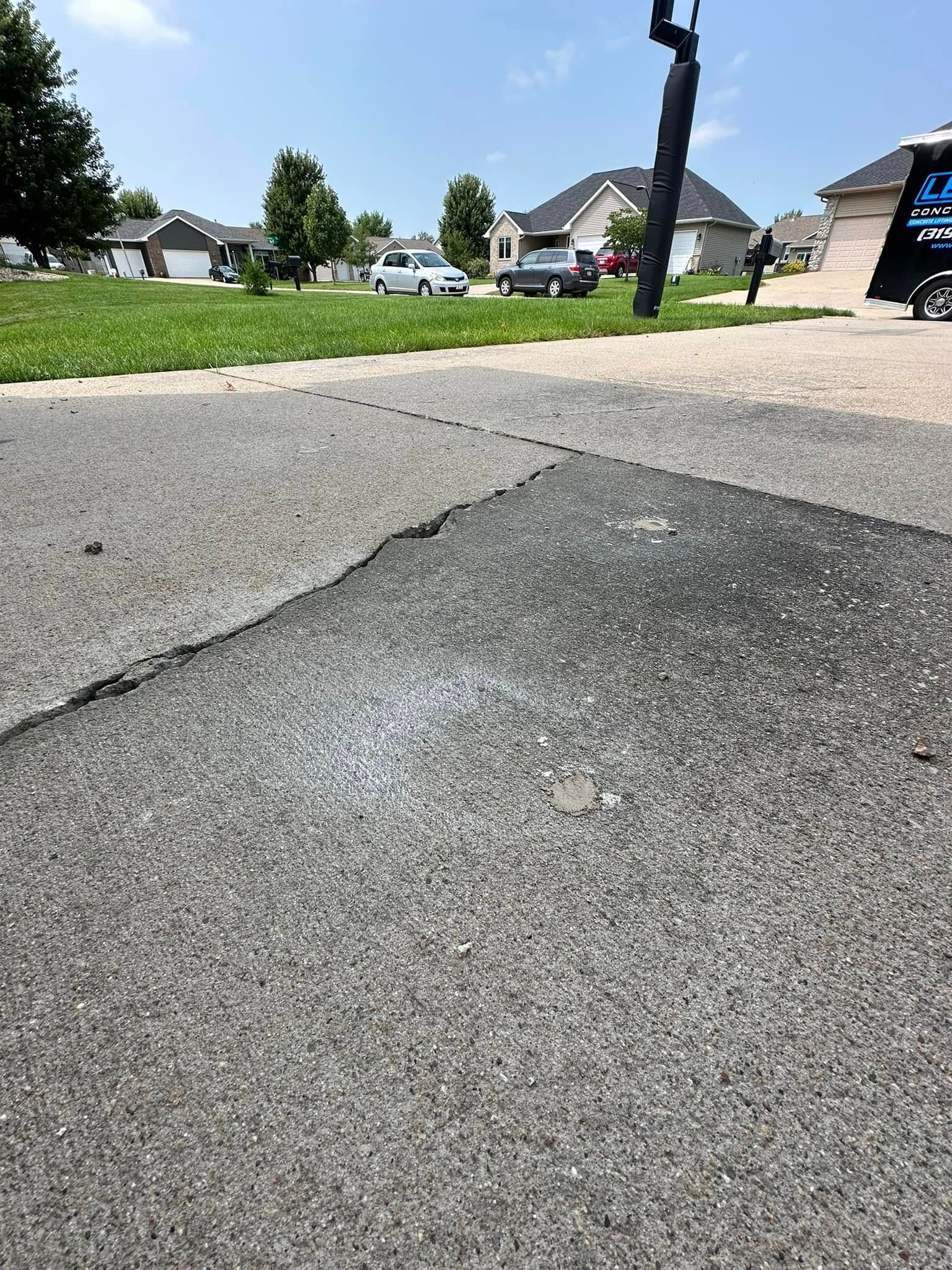 Close-up of asphalt road with a crack; grass, houses, cars, and blue sky in background.