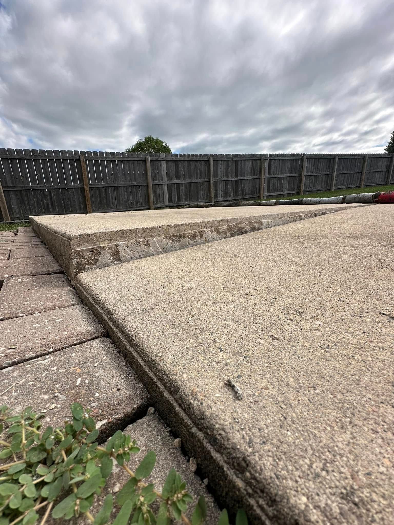 Concrete patio with edging next to a row of paving stones and a wooden fence under a cloudy sky.
