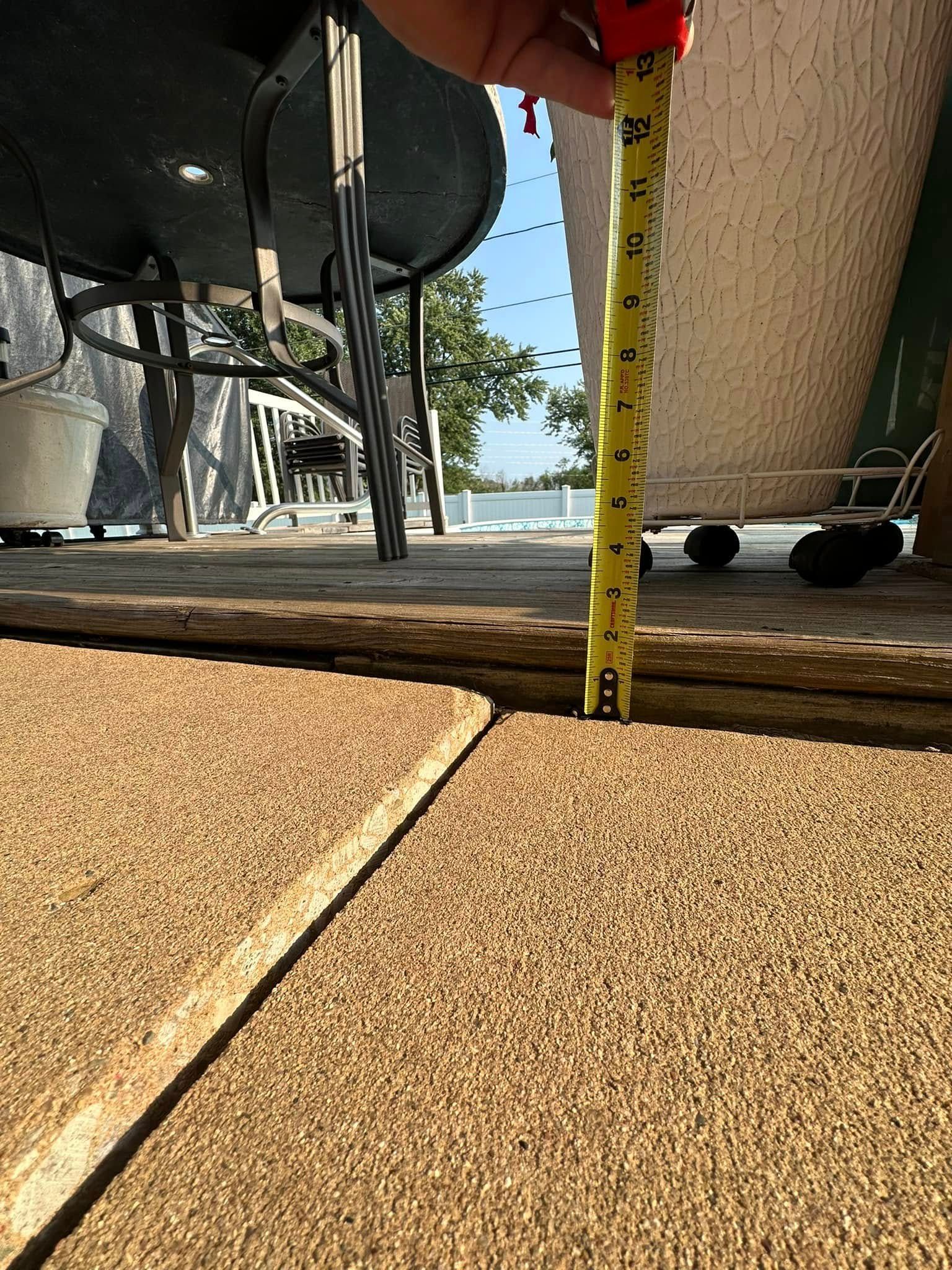 A hand holds a measuring tape in a large gap between two textured concrete slabs, outdoors.