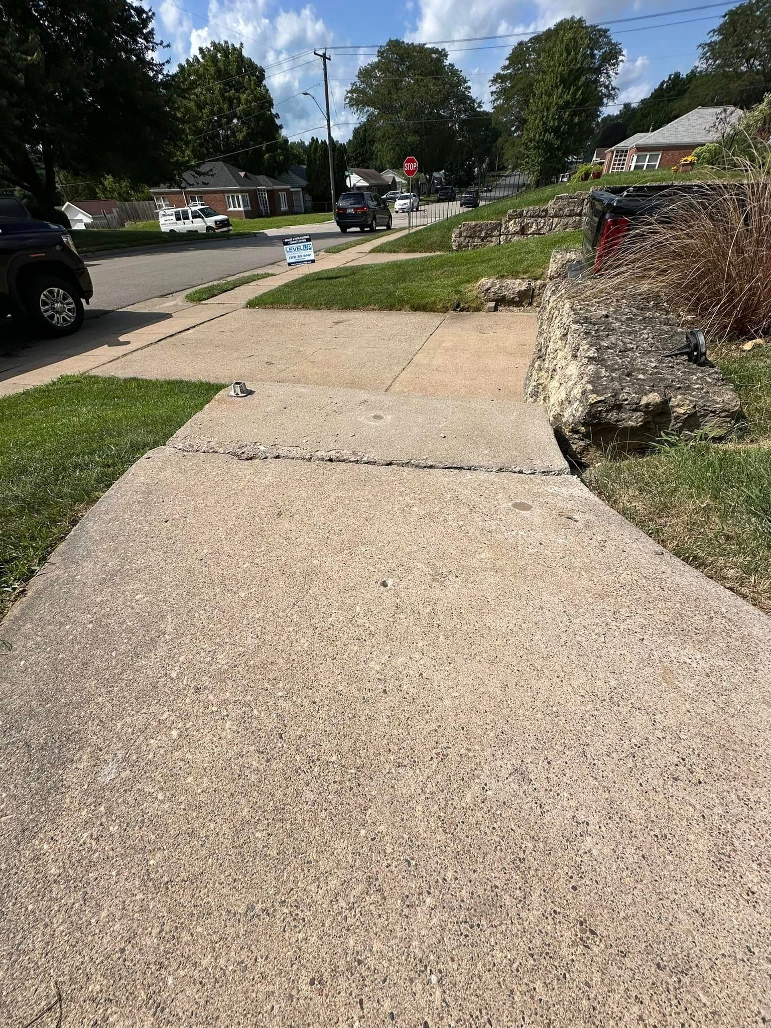 Sidewalk with cracked concrete, a grassy area, and a street with cars in a residential neighborhood.