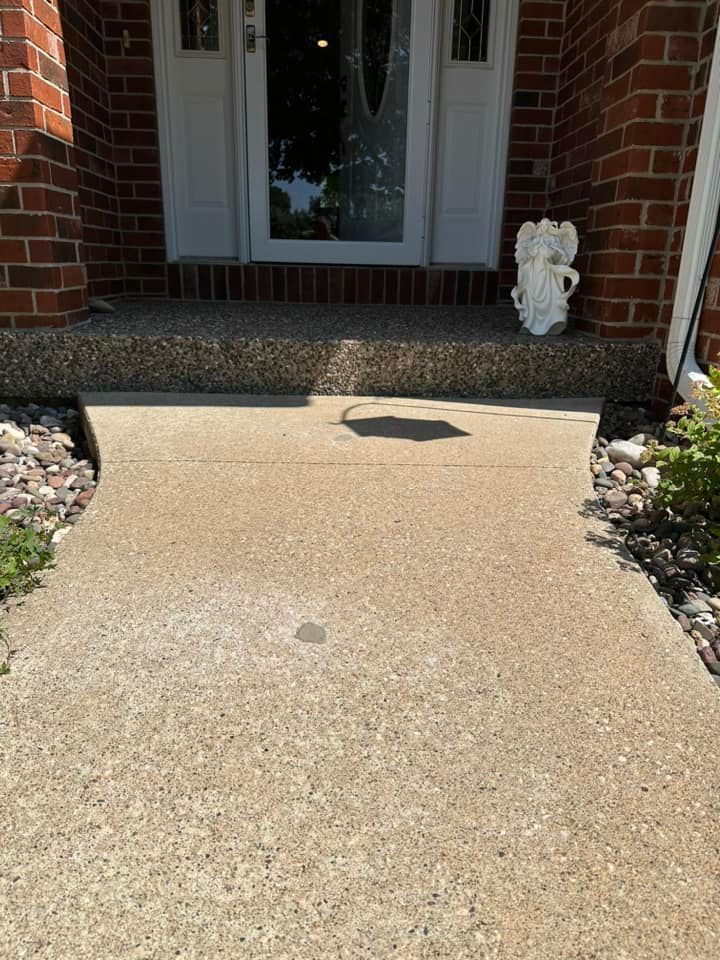 Concrete walkway leading to a front door with a small porch, flanked by flower beds and brick pillars.