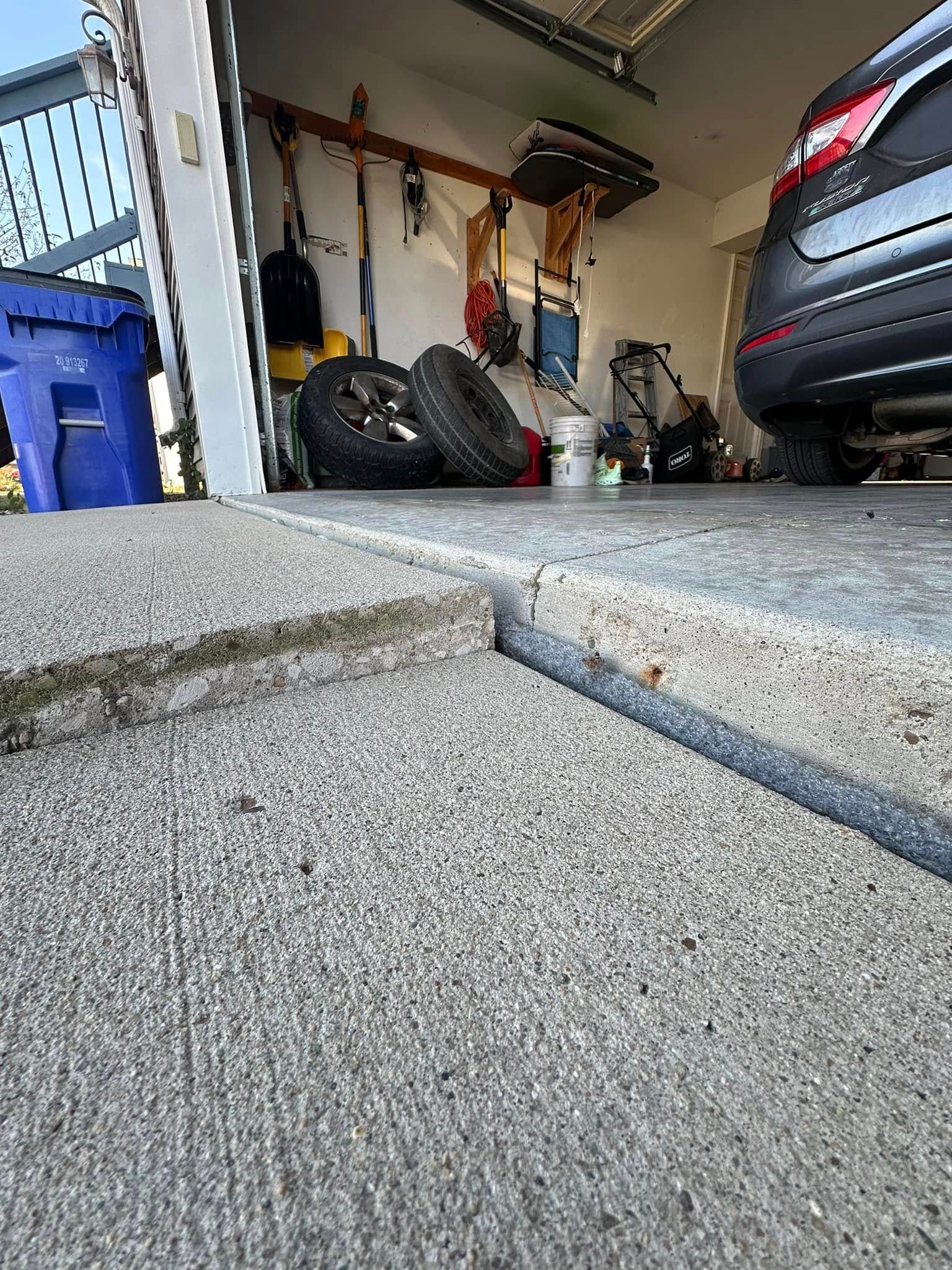 Cracked concrete driveway leading into a garage, with tires, tools, and a car inside.