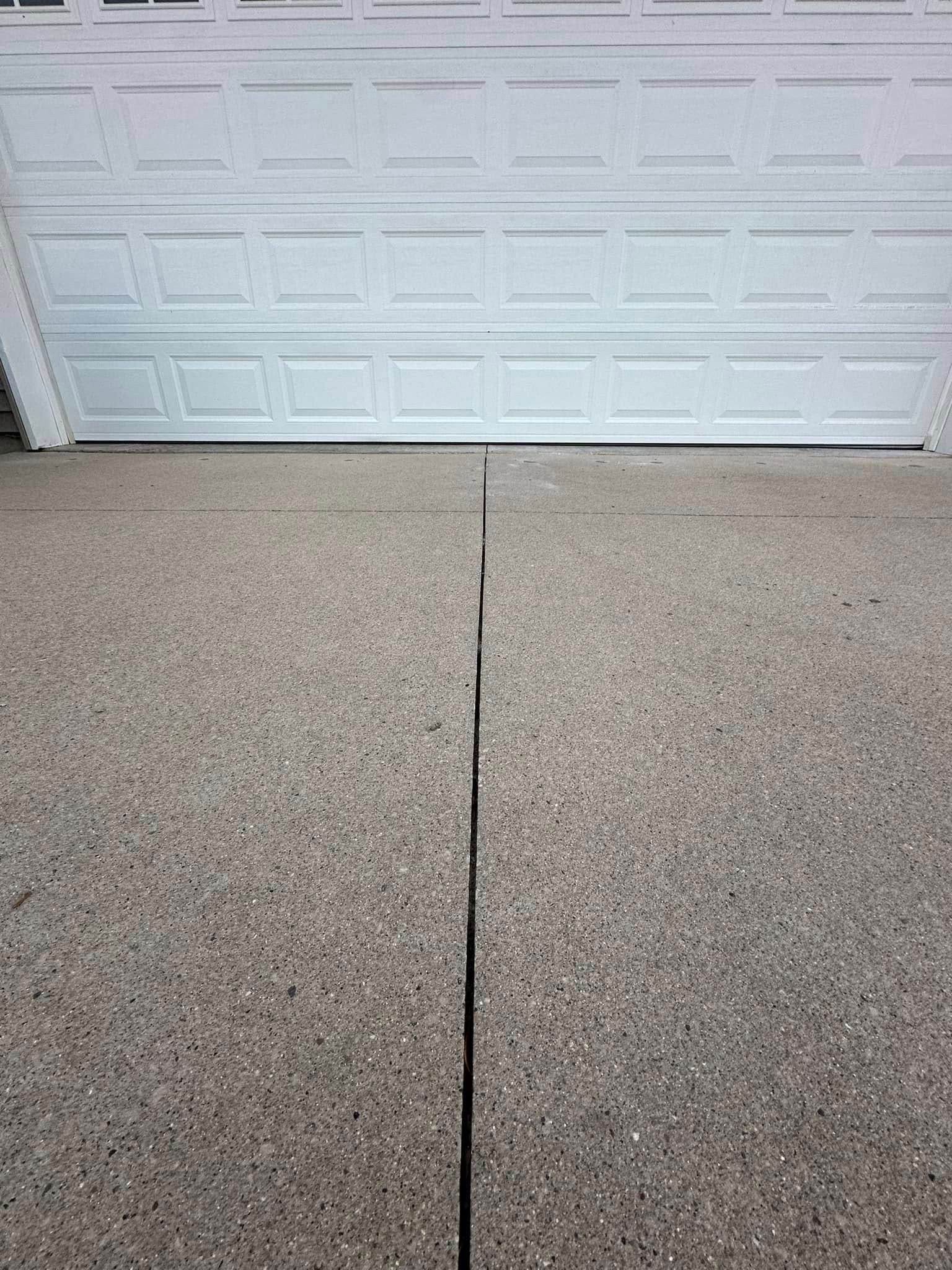 Concrete driveway with a large crack in the center, in front of a white garage door.