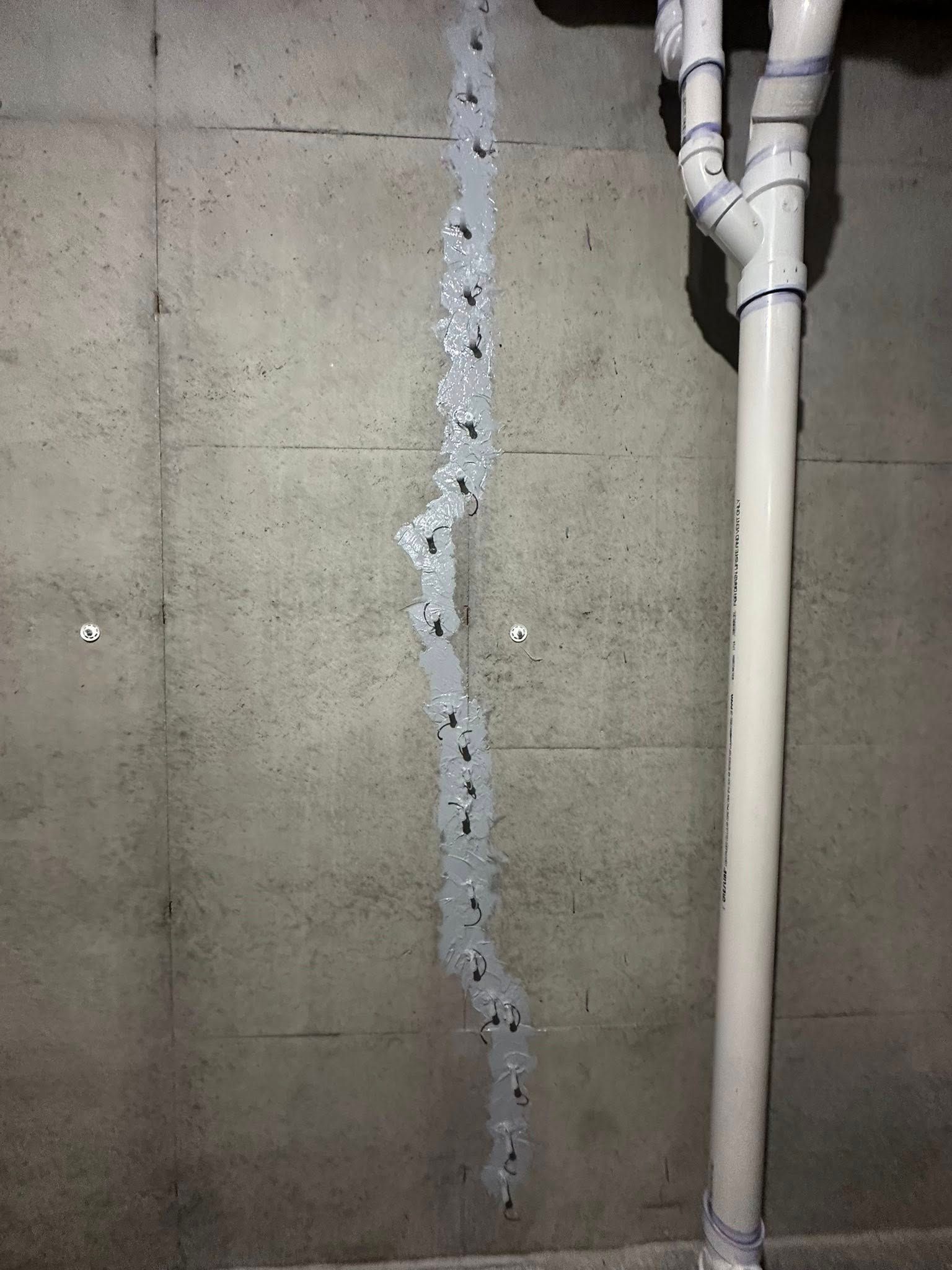 Concrete wall with a vertical crack filled with sealant, near plumbing pipes.
