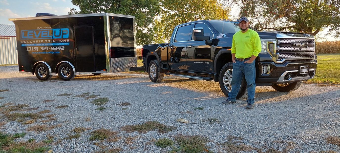 Man in neon shirt stands next to black truck and trailer. Gravel driveway, cornfield background.