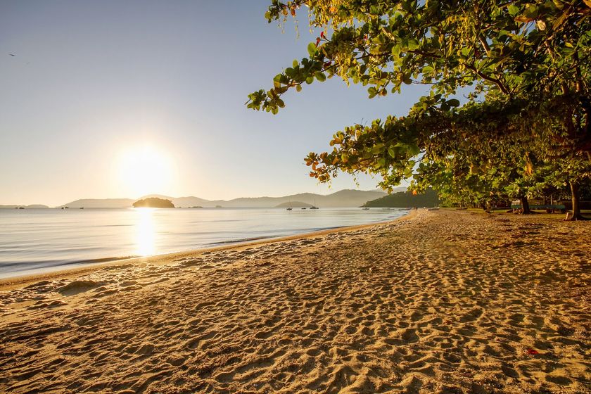 Praia de areia ao pôr do sol com águas calmas, ilhas distantes e árvores ao longo da costa.