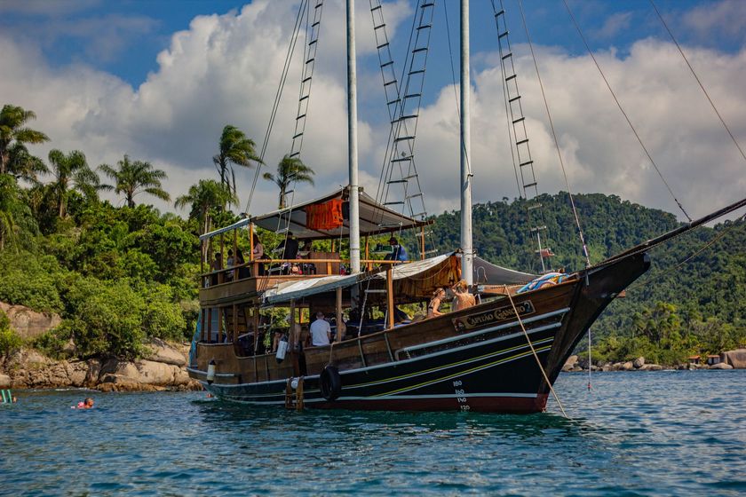 Barco à vela de madeira com passageiros em águas azuis perto de uma costa tropical e colinas verdejantes.