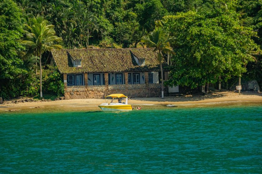 Cabana de praia com telhado de palha à beira de águas turquesa, com densas árvores verdes ao fundo e um caiaque amarelo na margem.
