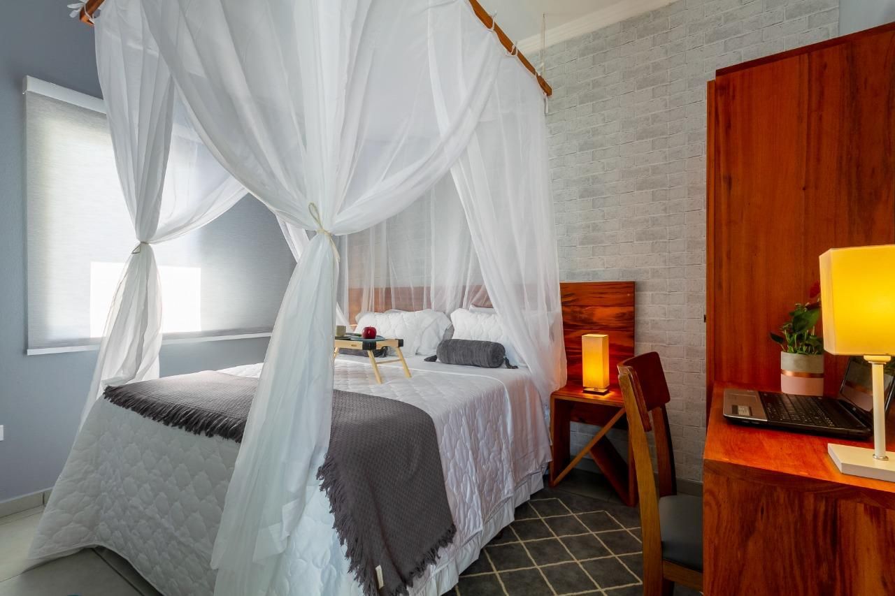 Quarto luminoso com cama de dossel branca, roupa de cama cinza, escrivaninha de madeira e luminária com luz quente.