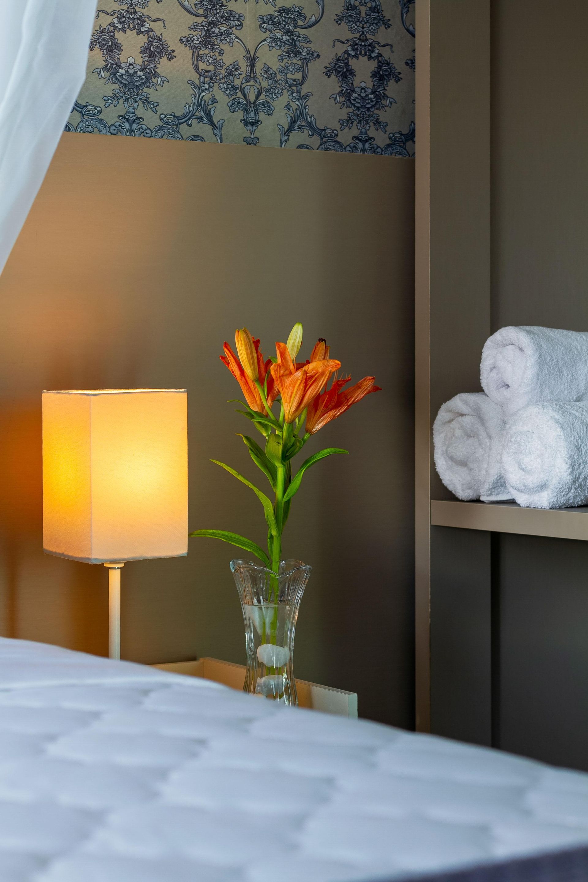 Quarto de hotel com abajur aceso ao lado da cama, flores laranjas em um vaso e uma escultura branca em uma prateleira.