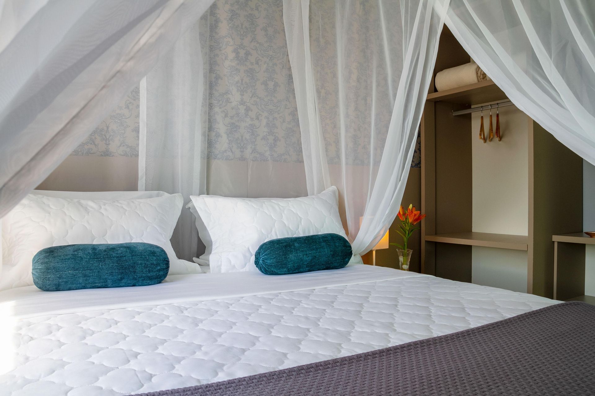 Cama com dossel, mosquiteiro branco, almofadas verde-azuladas e colcha branca em um quarto iluminado.