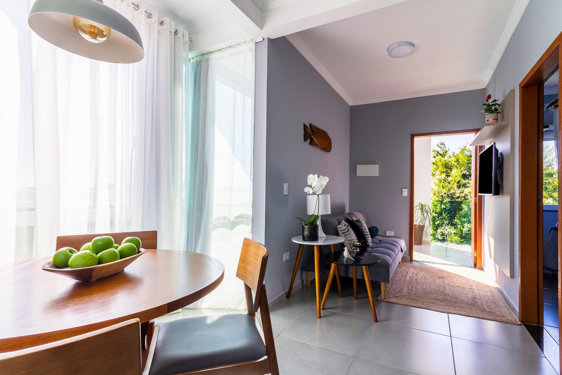 Sala de jantar iluminada com mesa redonda, maçãs verdes, cadeiras cinza e porta aberta para um pátio.