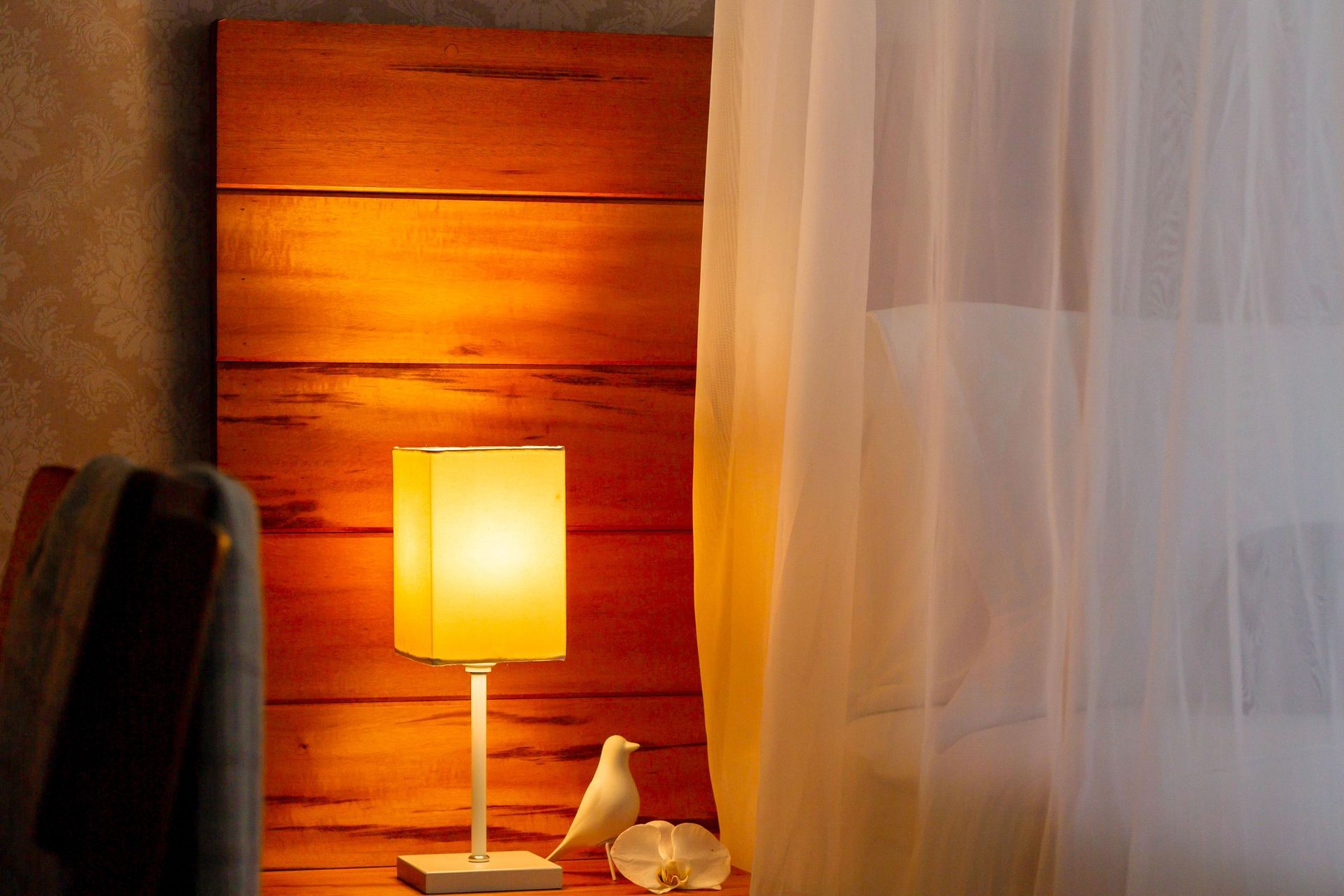 Abajur aconchegante iluminando a cabeceira ao lado de uma estatueta de pássaro branco e cortinas translúcidas em um quarto acolhedor.