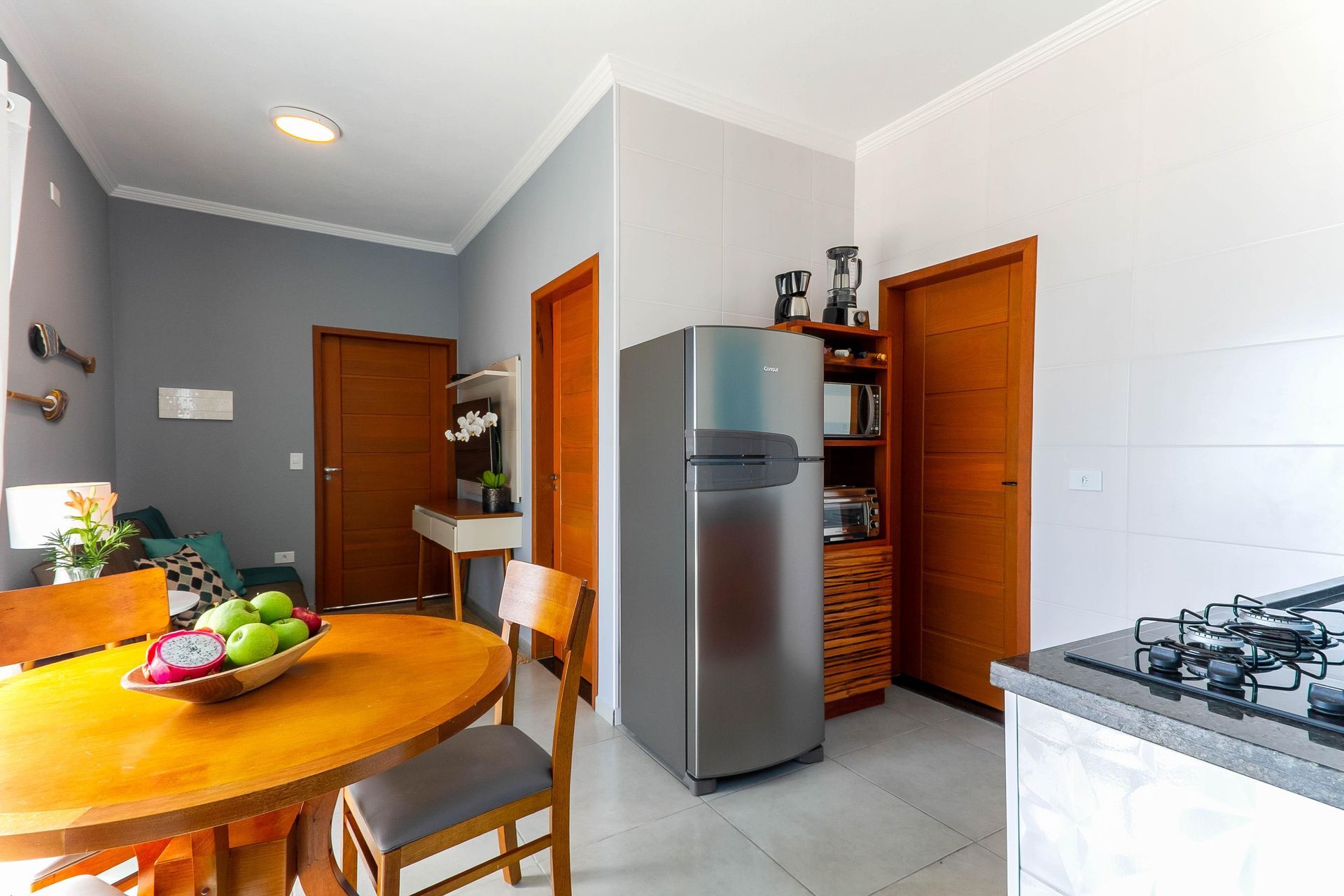 Cozinha iluminada com mesa redonda de madeira, geladeira, fogão a gás e fruteira perto das portas de madeira.