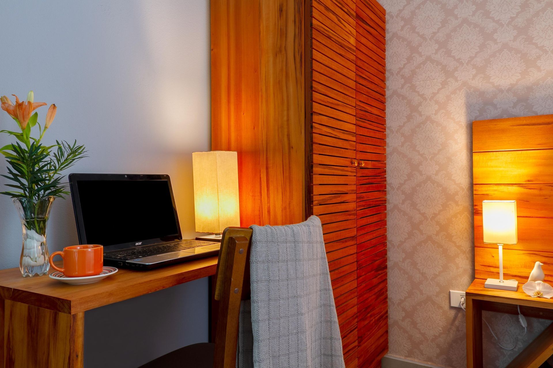 Espaço de trabalho aconchegante no quarto com laptop, caneca laranja, luminária e cadeira coberta com uma toalha cinza.