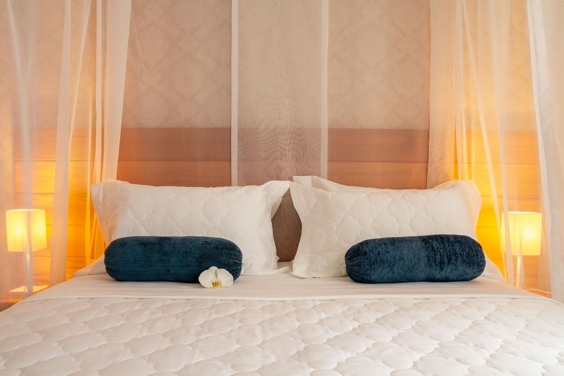 Cama aconchegante com lençóis brancos, travesseiros azuis, cortinas de dossel transparentes e abajures com luz quente.