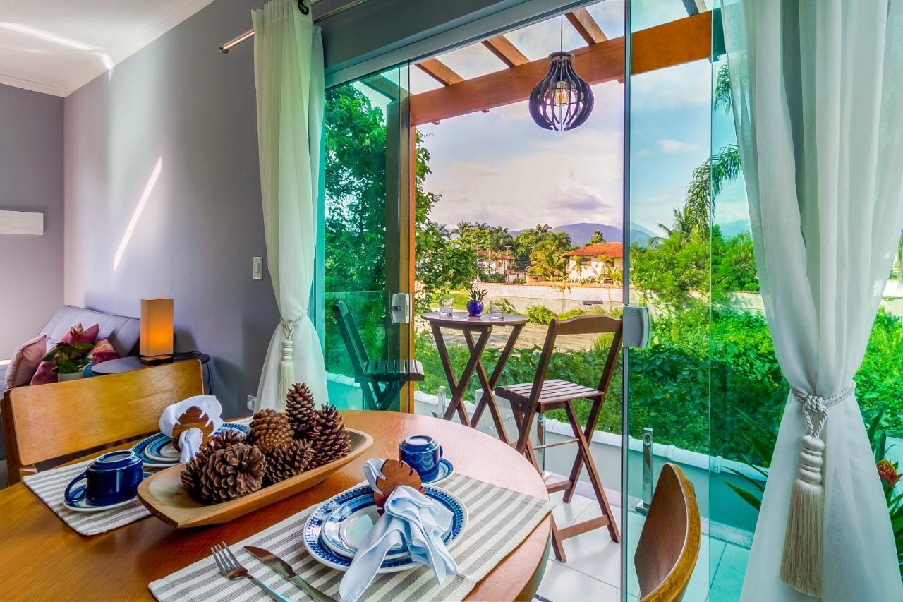 Mesa de jantar iluminada junto às portas abertas da varanda, com cadeiras de madeira, guardanapos azuis e vista para um jardim tropical.