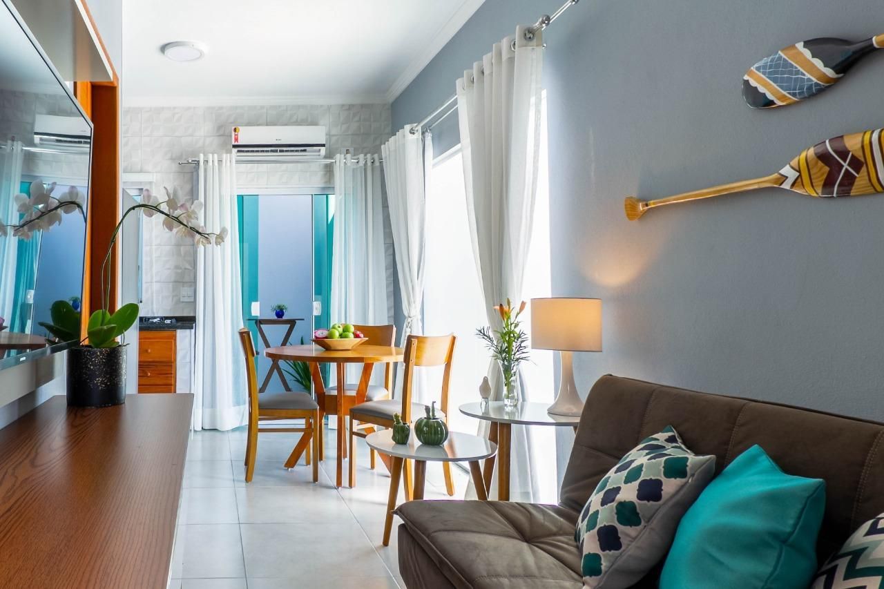 Sala de estar estreita e iluminada com sofá, mesa de jantar, detalhes em azul e luz solar entrando pelas cortinas translúcidas.