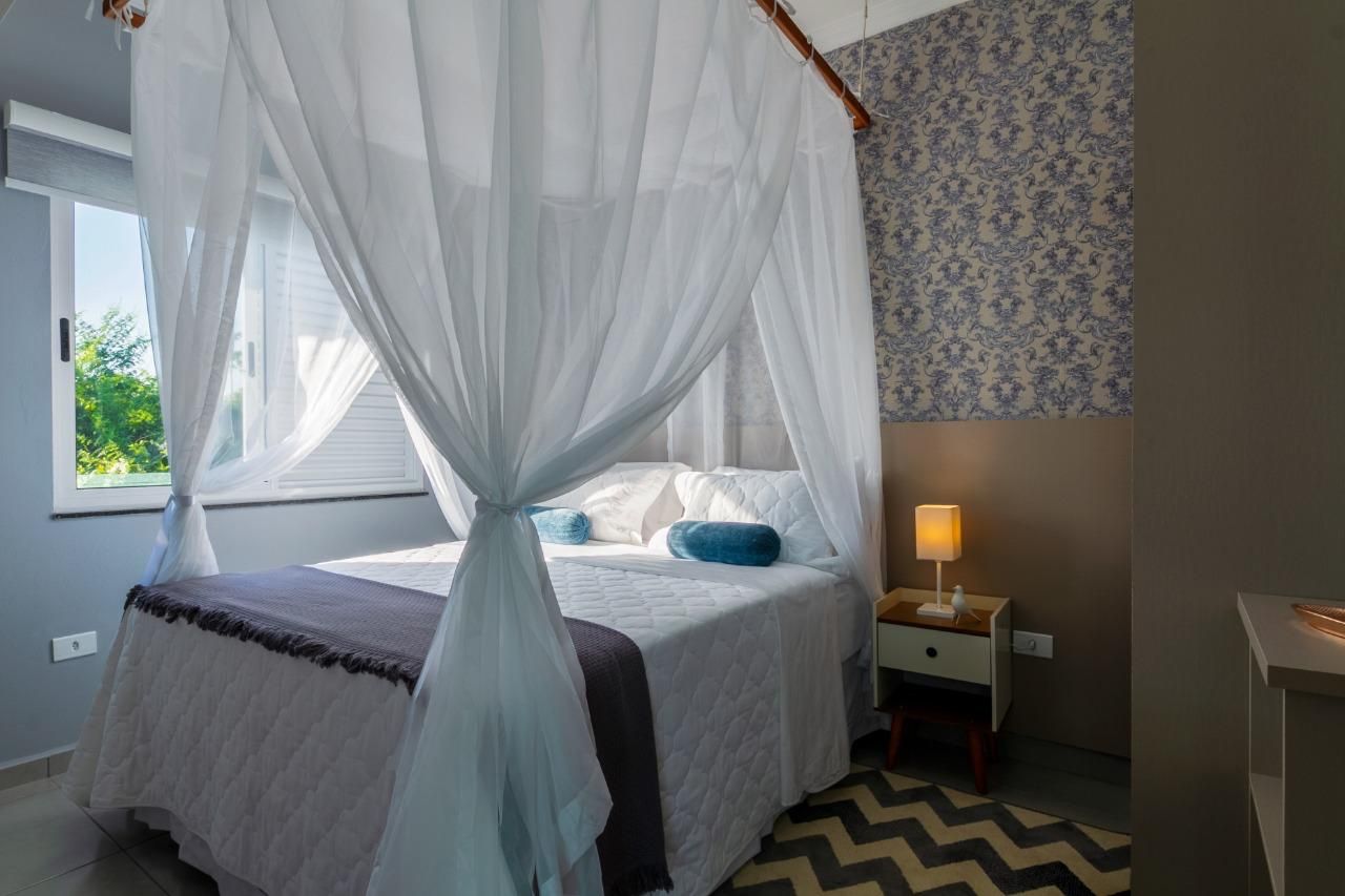 Quarto luminoso com cama de dossel, cortinas brancas com dossel, abajur e papel de parede estampado.