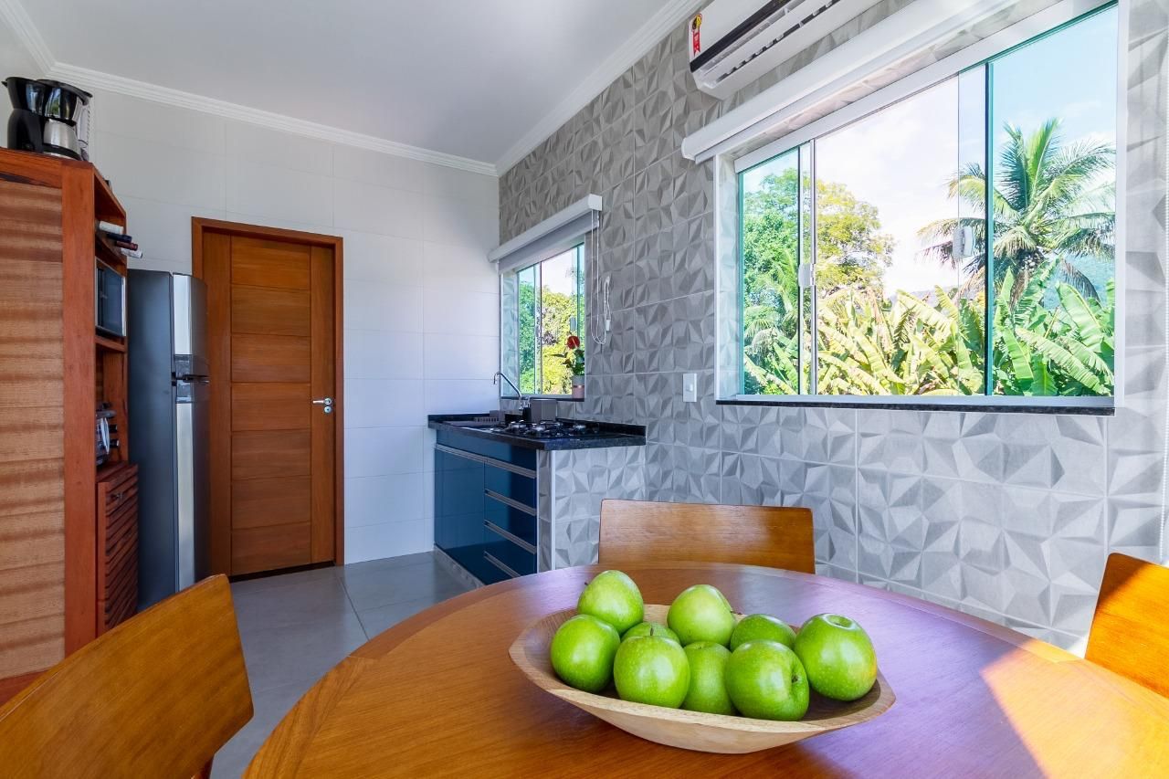 Sala de jantar iluminada com mesa de madeira, fruteira verde, cômoda azul e grande janela com vista tropical.