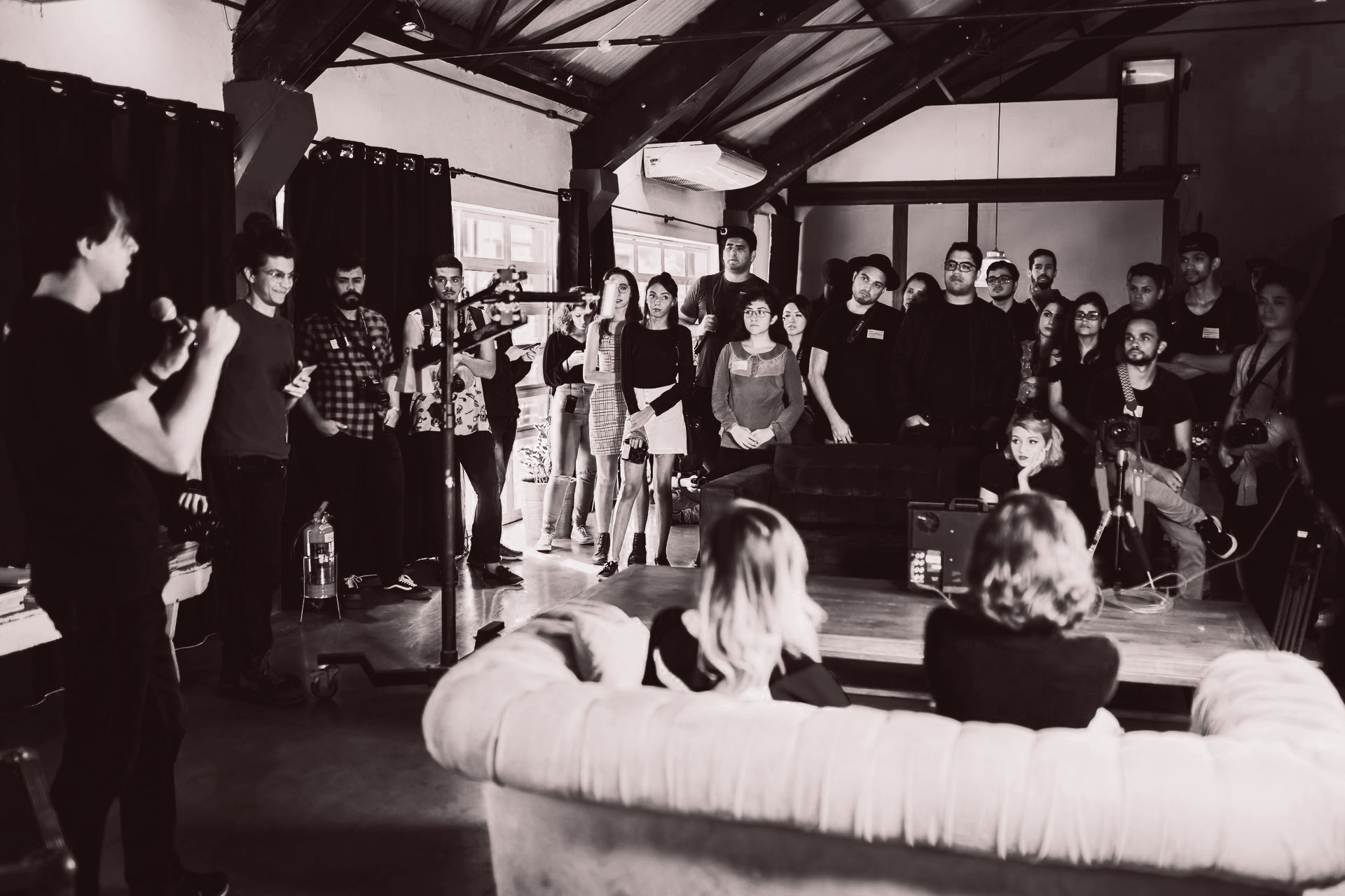 People gathered at a venue, likely a performance. Two women sit on a couch as others stand around, watching.