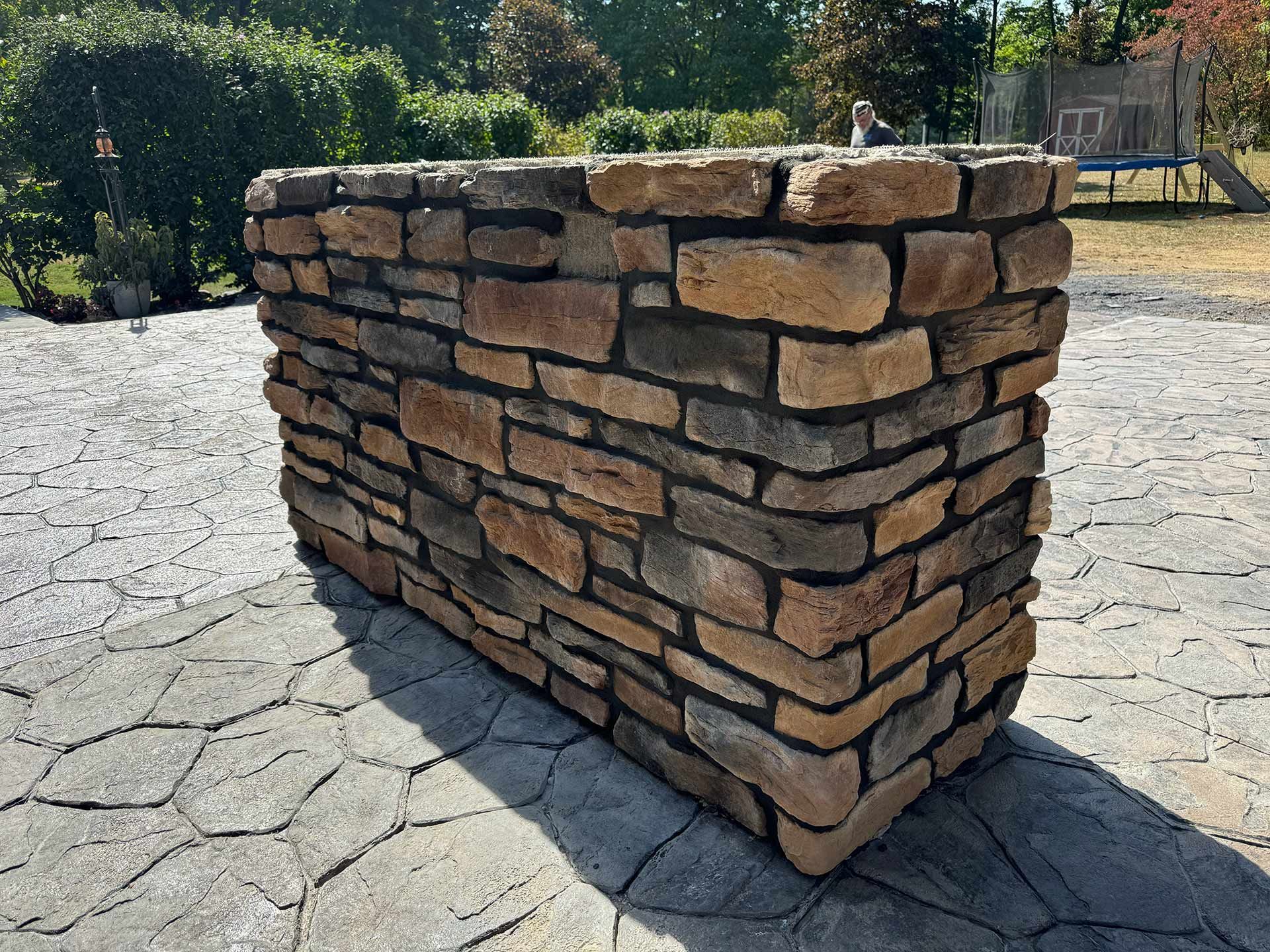 Brick planter, rectangular, made of various brown and tan bricks, set on a patterned concrete surface, outdoor setting.