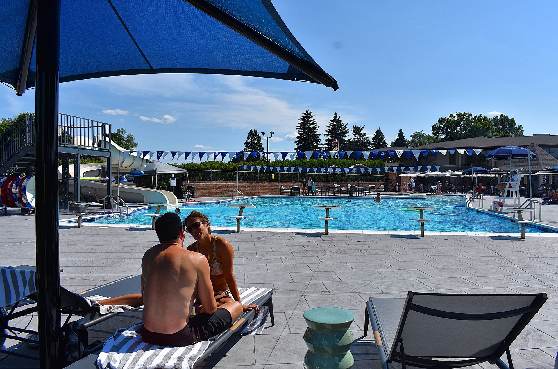 Two people sitting by a blue swimming pool under a canopy, with lounge chairs and umbrellas nearby.