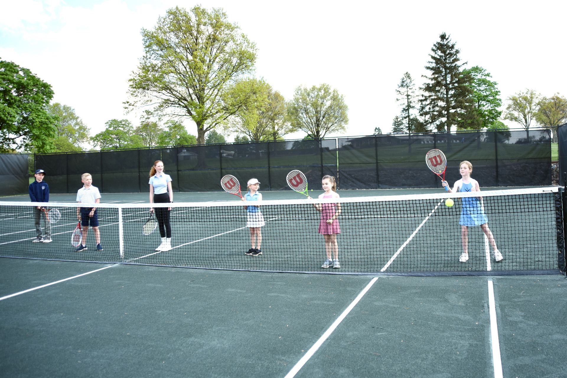 Children standing on a tennis court behind a net, holding rackets near the baseline.