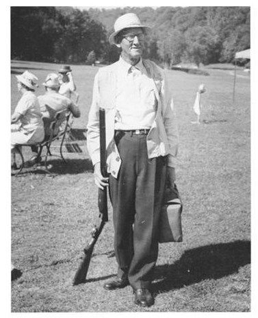 Black-and-white photo of a man in a hat holding a rifle and bag on a golf course.