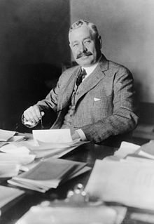 Man in suit seated at a desk with papers, looking toward the camera