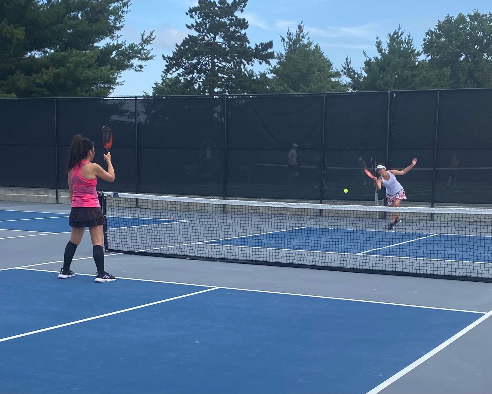 Two people play tennis on a blue court, one serving near the net and one returning at the far end.