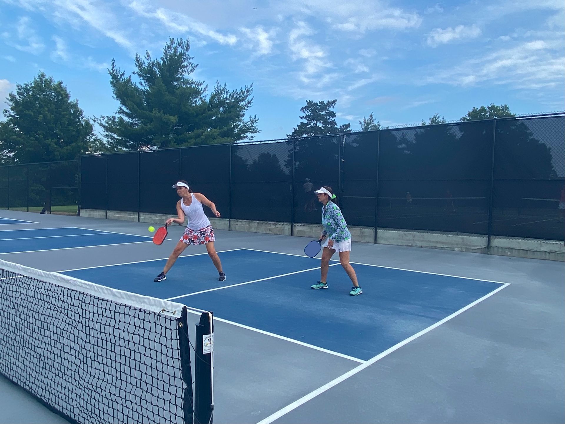 Two players rally on a blue outdoor tennis court beside a black windscreen.