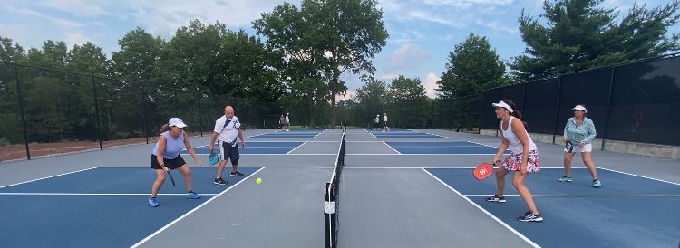 Four people playing pickleball on outdoor blue courts with a net and trees in the background