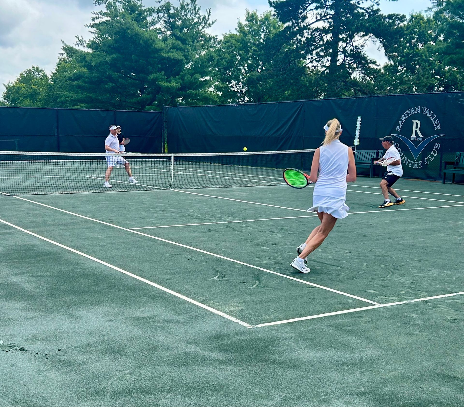 Three people playing tennis on an outdoor court with a green fence and trees behind them