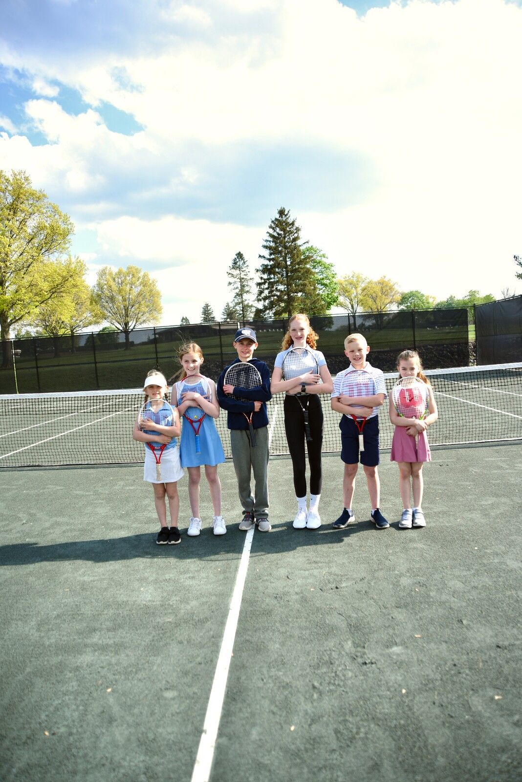 Six children standing on a tennis court, holding trophies, with a net and trees in the background.