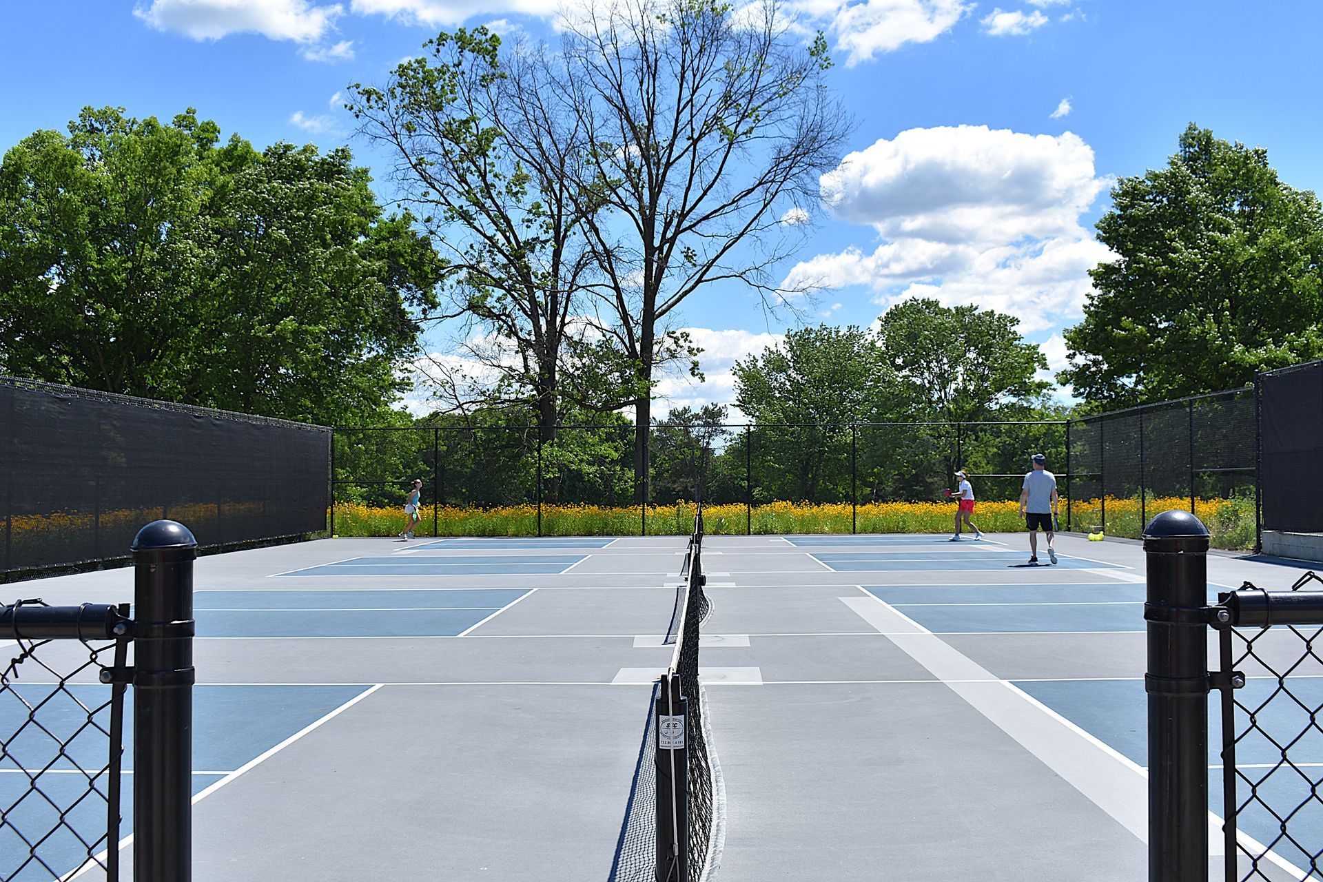 Outdoor tennis court with blue and gray surface, a few players, and trees under a partly cloudy sky