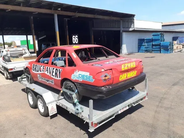 Red race car on a trailer outside a garage, with sponsor decals and a spare tire mounted on the side — Ezyas Car Wash NT in Yarrawonga, NT