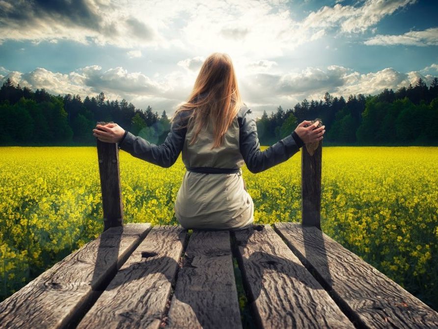 Person sitting on a wooden dock overlooking a bright yellow field and cloudy sky