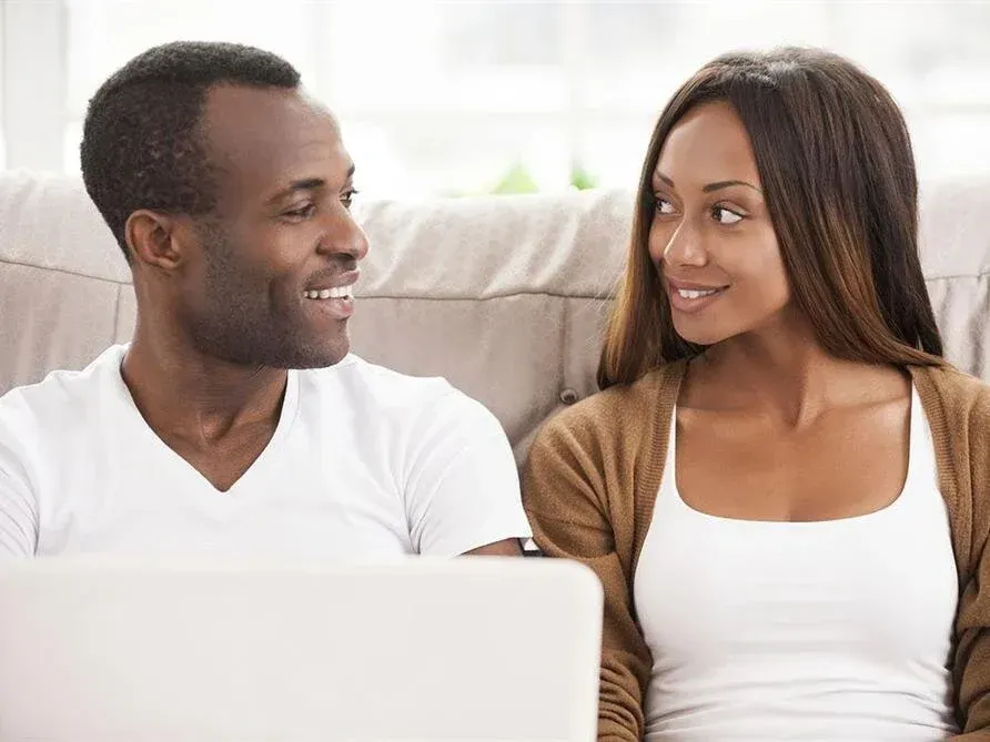 Two people smiling at each other on a couch with a laptop in the foreground.