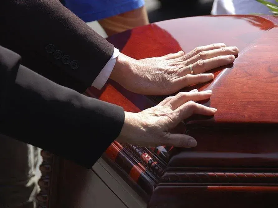 Hands resting on a polished red casket at a funeral