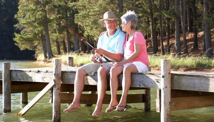 Two people sit on a wooden dock by a lake, one wearing a hat and holding a fishing rod.
