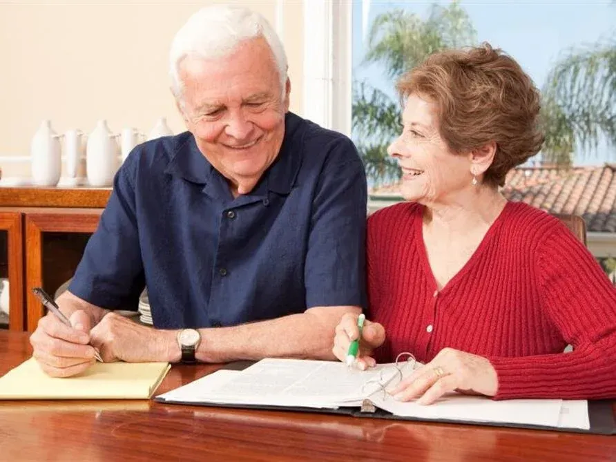 Two smiling people sit at a table, writing in a notebook with a pen and green marker.
