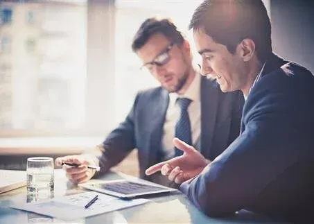 Two businesspeople discussing documents at a desk with a laptop and coffee in a bright office