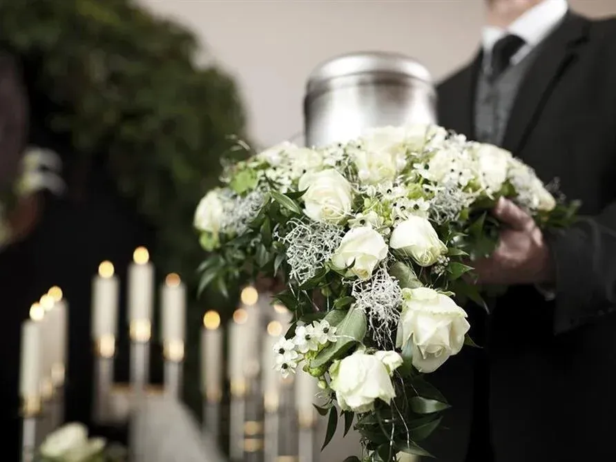White floral funeral spray beside lit candles, held by a person in a dark suit