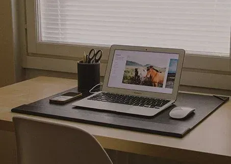 Laptop on a desk by a window, with a mouse, phone, pen cup, and notebook.